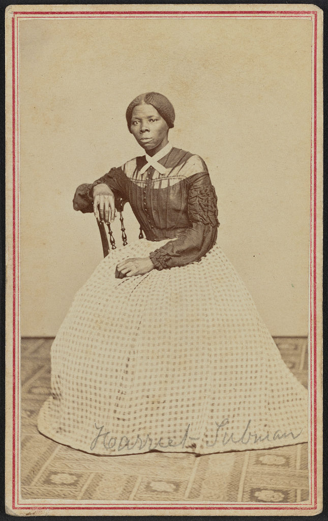 Photograph shows Harriet Tubman (1822-1913) at midlife. She is seated, turned toward the left. One hand rests on the back of a wooden chair, another rests in her lap.