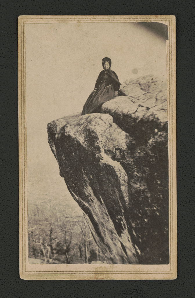 Photograph shows nurse Grace sitting on a rocky ledge. She is wearing dark clothes and a dark bonnet.