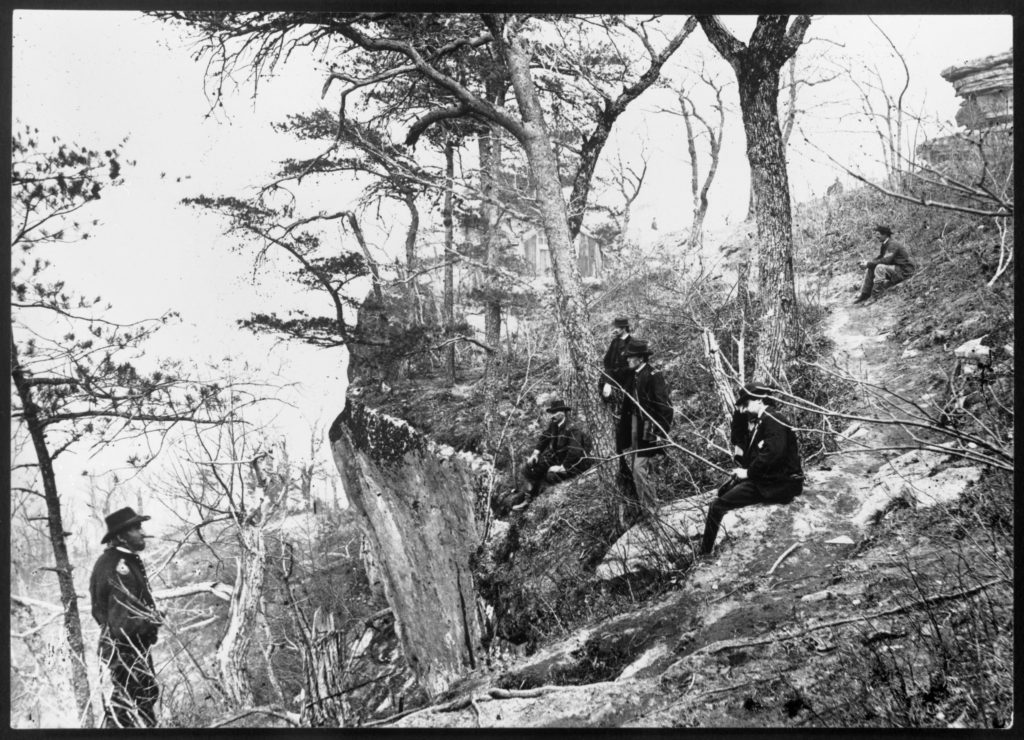 General Grant, cigar in his mouth, stands near a precipice on a hillside. At least five other men, several of them officers, are also on the hillside. Grant looks to the photo's right, while the other men are looking toward the left of the photo, likely toward a battlefield.